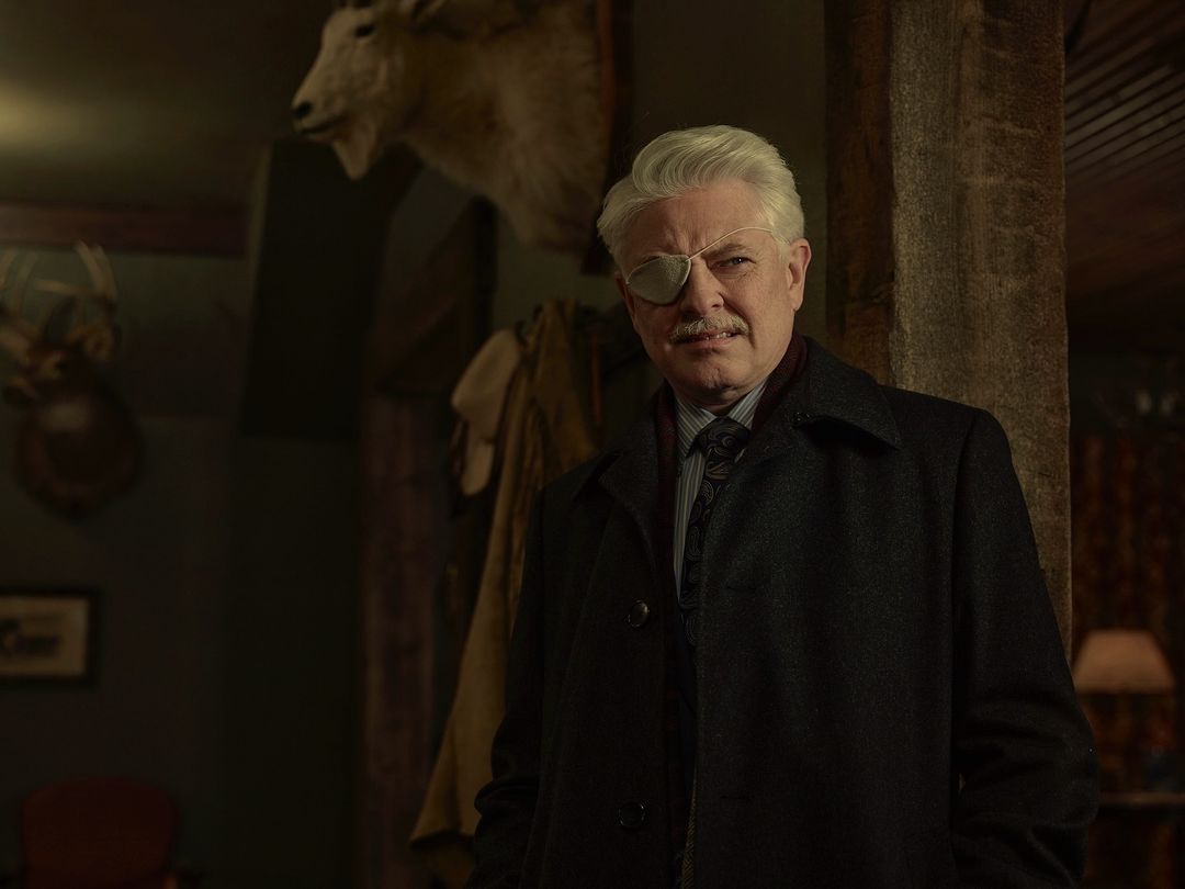 Older gentleman in dark coat and eye patch standing indoors with animal taxidermy displayed in the background, portrait orientation.