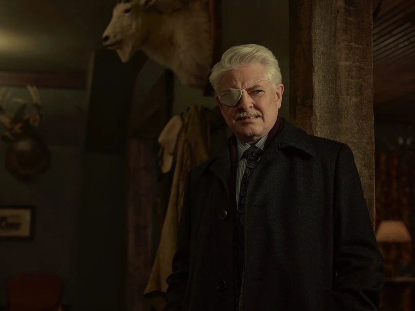 Older gentleman in dark coat and eye patch standing indoors with animal taxidermy displayed in the background, portrait orientation.