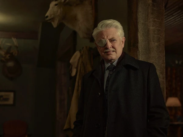 Older gentleman in dark coat and eye patch standing indoors with animal taxidermy displayed in the background, portrait orientation.