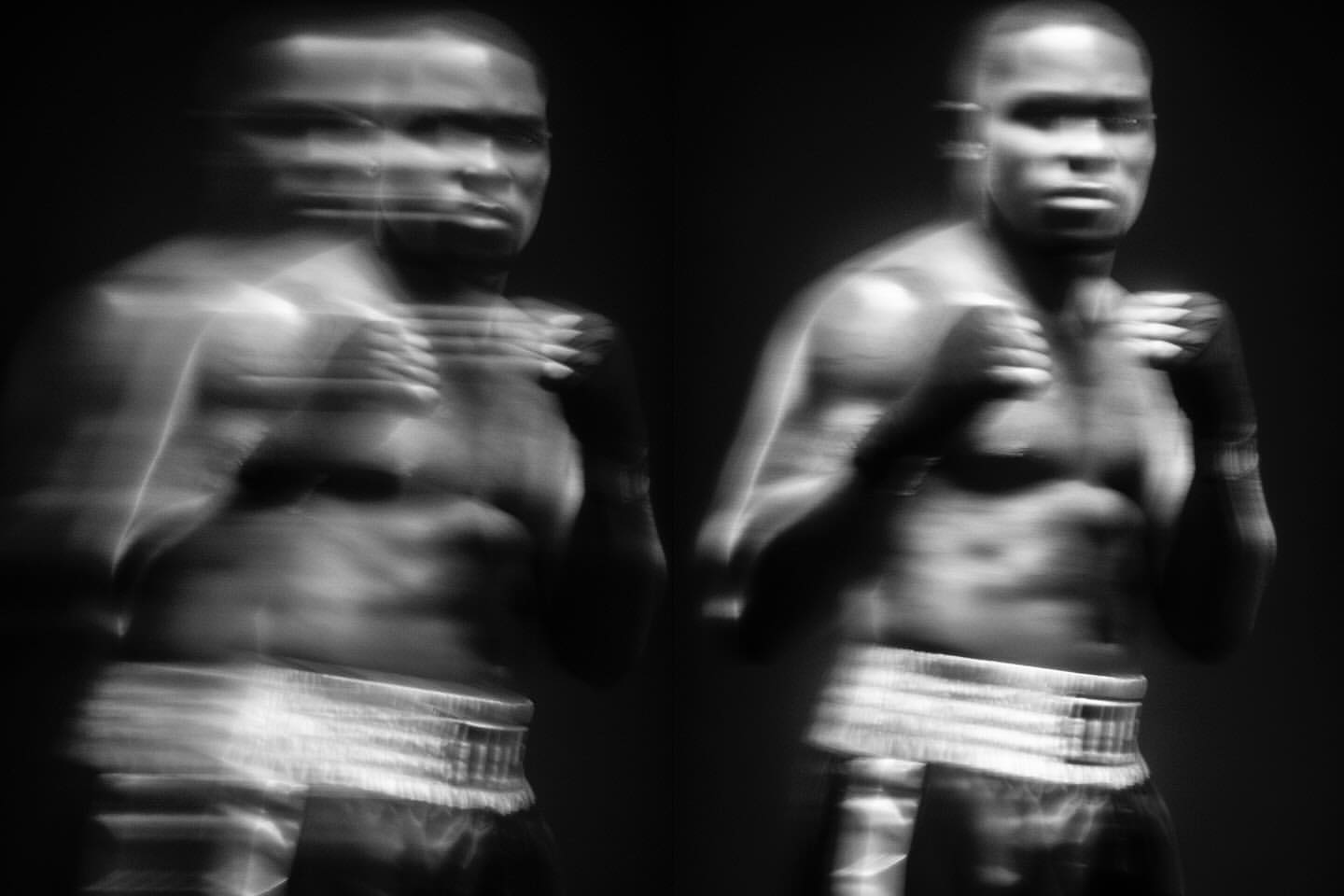 Black and white portrait of a muscular African American man in boxing stance with motion blur, set against a dark, nondescript background.