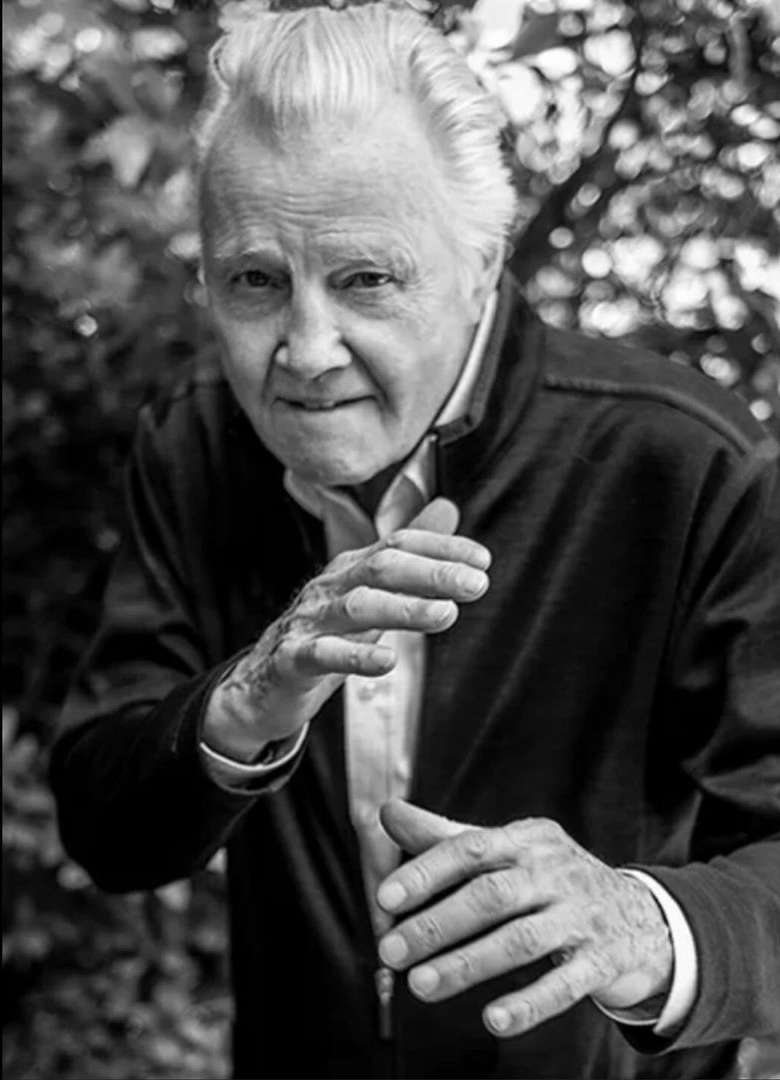 Black and white portrait of an elderly man in a jacket with his hands slightly raised in front of a blurred leafy background.