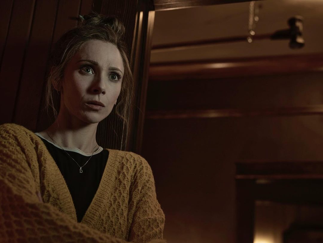 Woman in a yellow cardigan with a concerned expression, inside a dimly lit interior setting with dark wood paneling visible.