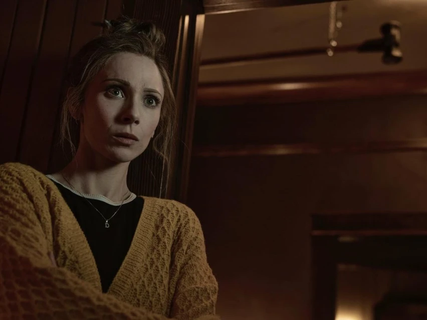 Woman in a yellow cardigan with a concerned expression, inside a dimly lit interior setting with dark wood paneling visible.