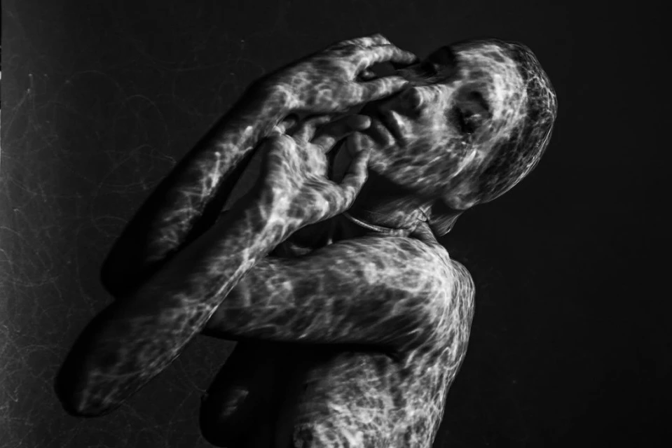 Monochrome portrait of an adult with textured lighting effects on the face and body, set against a dark background in a studio setting.
