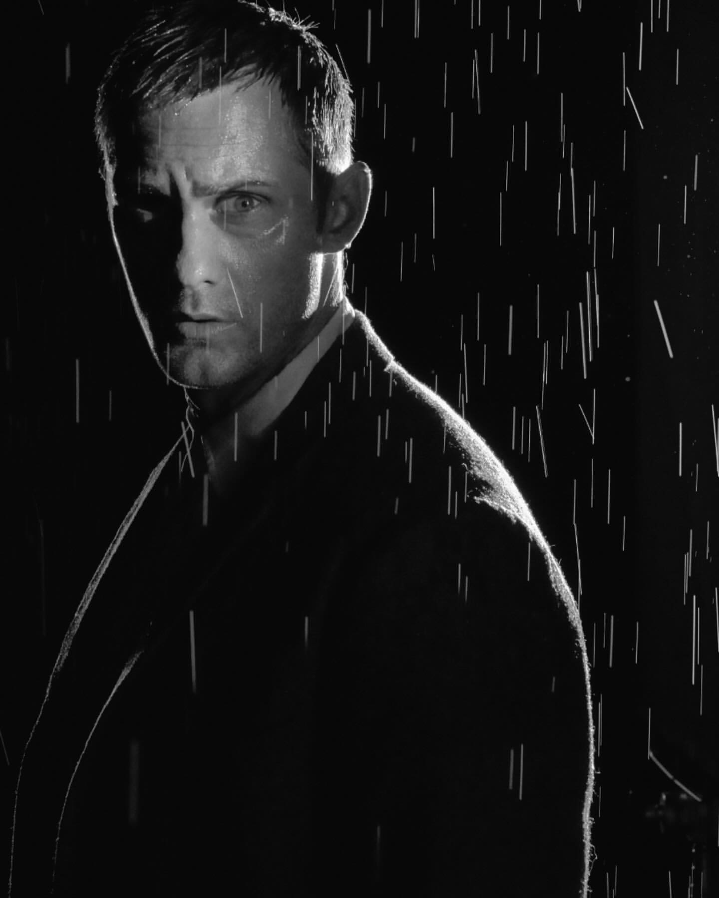 Monochrome portrait of an adult male in a suit standing in rain, dark background, dramatic lighting, creating a cinematic mood.