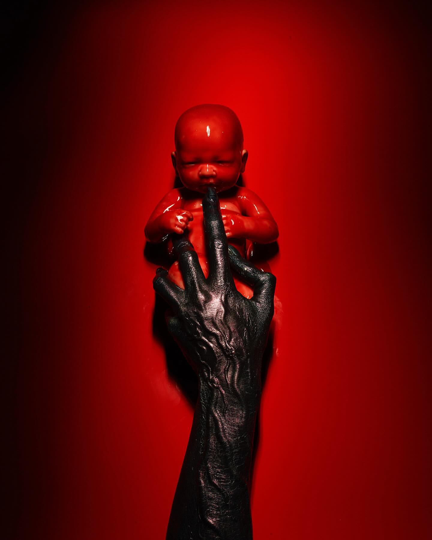 An eerie, red-toned image features a red baby doll held by a black-painted arm and hand, set against a matching red background. Dark mood.
