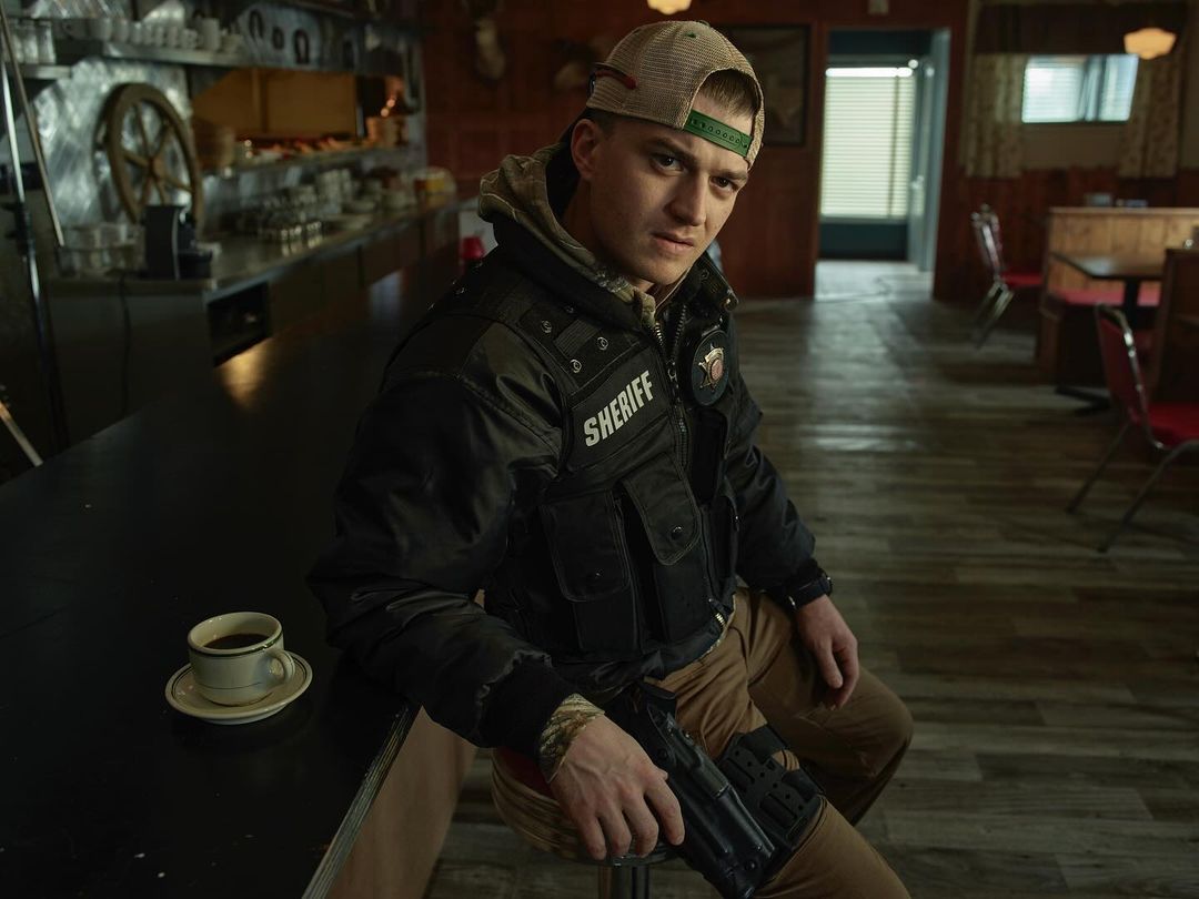 Indoor shot of a man in a uniform sitting at a counter with a coffee cup and gun, set against a dimly lit diner background.