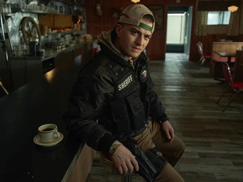 Indoor shot of a man in a uniform sitting at a counter with a coffee cup and gun, set against a dimly lit diner background.