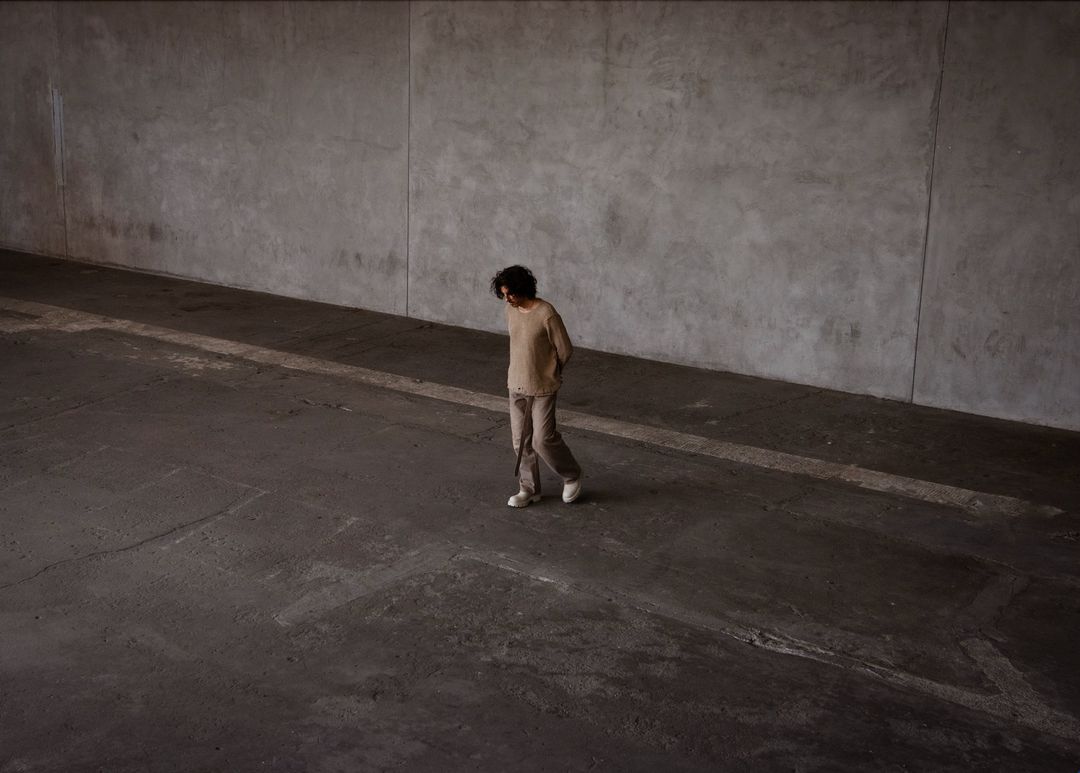 An adult walks in a large concrete space, wearing a beige sweater, pants and white shoes, looking down with natural lighting.