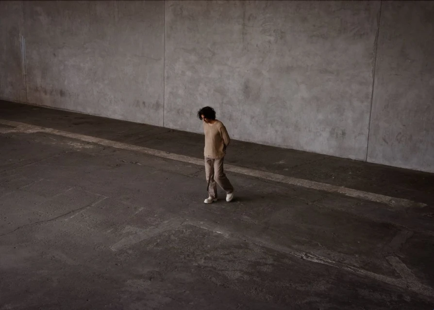 An adult walks in a large concrete space, wearing a beige sweater, pants and white shoes, looking down with natural lighting.