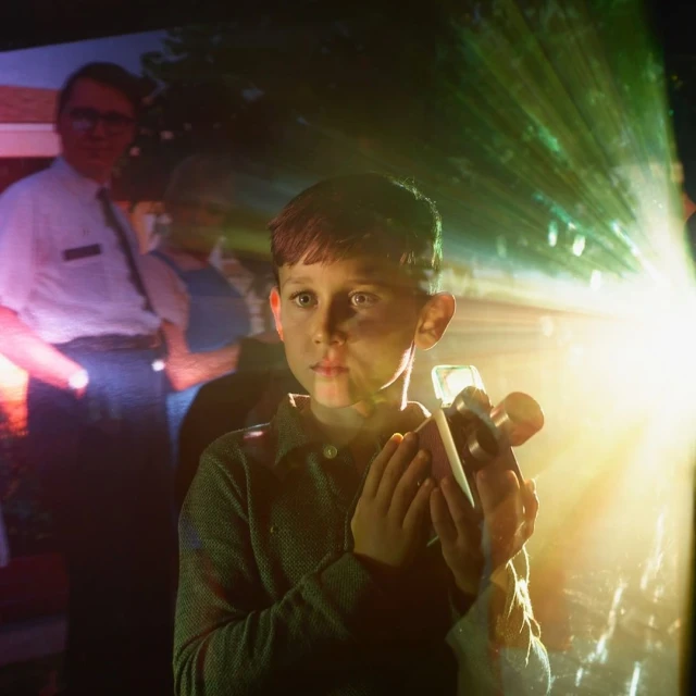 A young boy holds a toy projector in a darkened room, beams of light emitting from it, while adults stand blurred in the background.