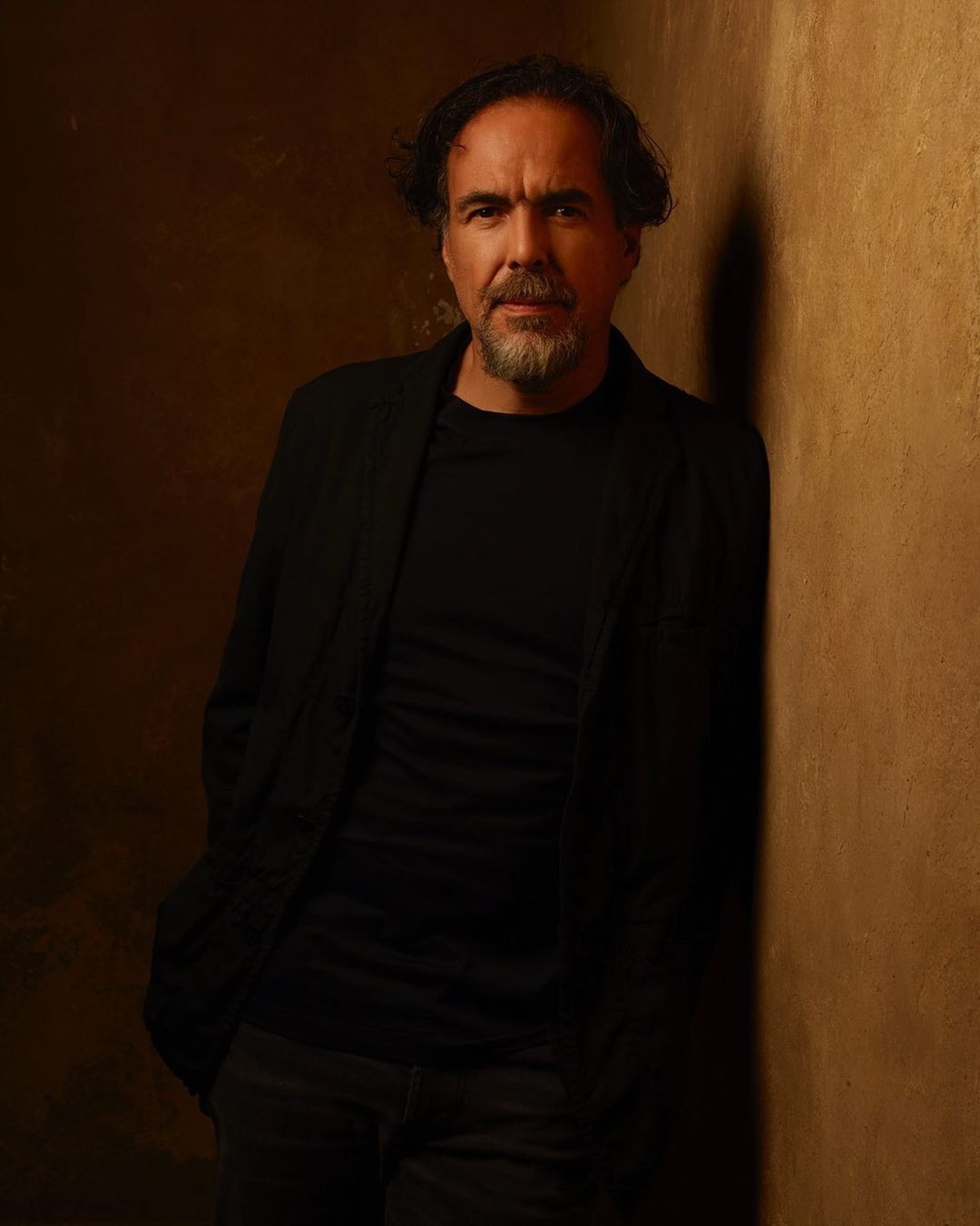Portrait of a middle-aged adult male with a beard wearing a black shirt and jacket against a tan textured wall in a photo studio.