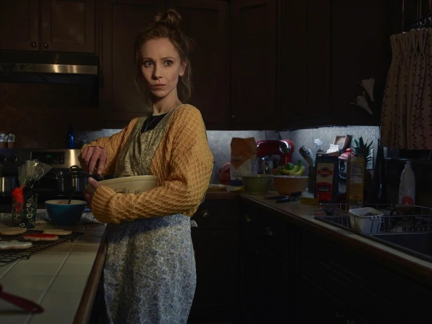 Woman in kitchen mixes ingredients in bowl while standing at counter with various cooking utensils and appliances, illuminated by dim lighting.