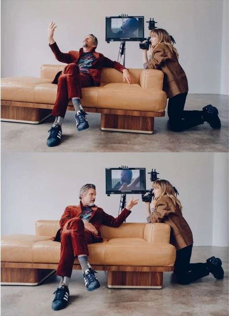 Man in a red suit sits on a tan leather couch while a woman kneels and films him with video equipment in a minimalist studio space.