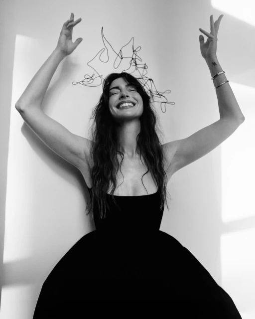 A monochrome portrait of a smiling woman wearing a black dress with arms raised, accessorized with wire art and silver bracelets.