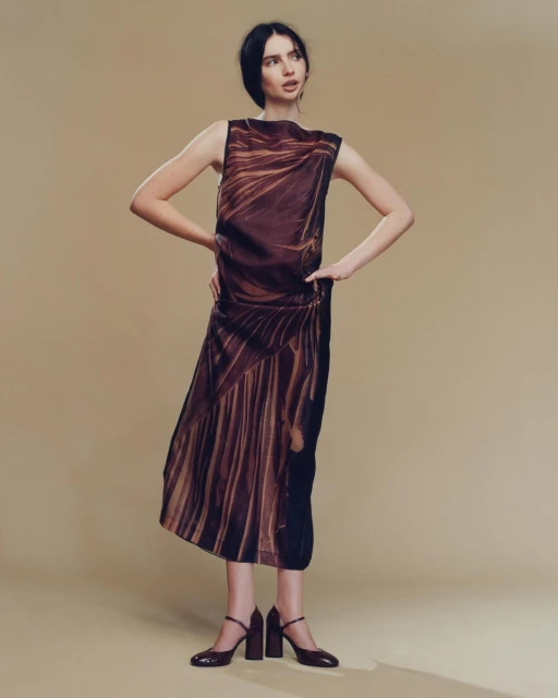 Brunette fashion model wears a brown patterned dress with mary jane heels against a light brown backdrop in a studio setting.