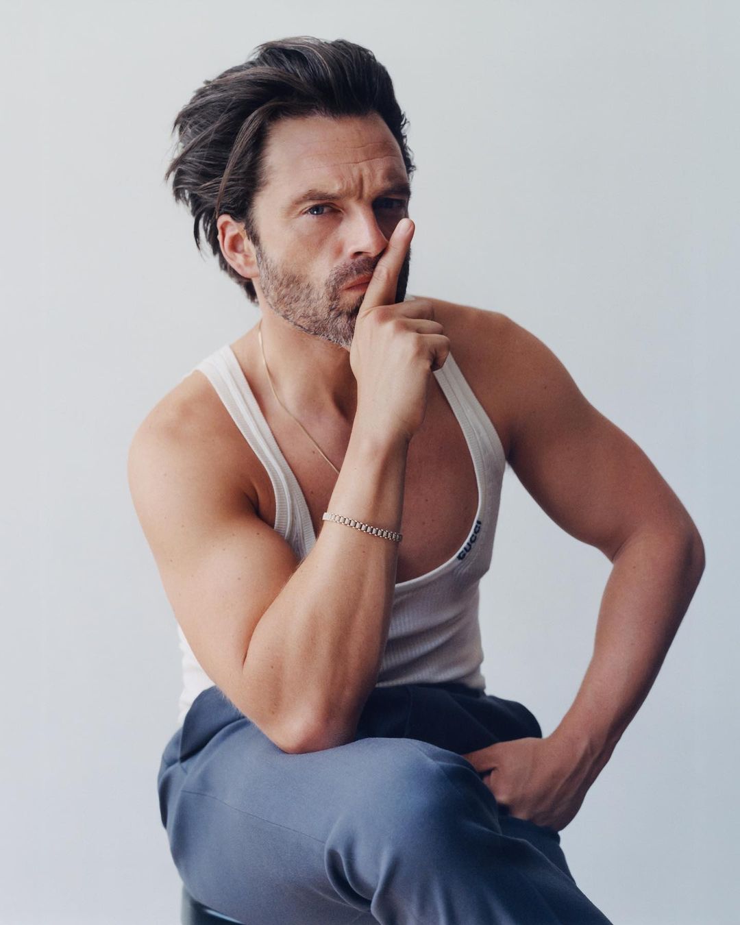 A brunette male model in a white tank top and blue sweatpants sits in a photo studio with his finger raised to his lips.