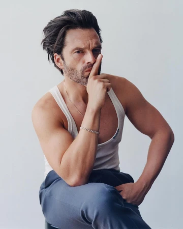 A brunette male model in a white tank top and blue sweatpants sits in a photo studio with his finger raised to his lips.
