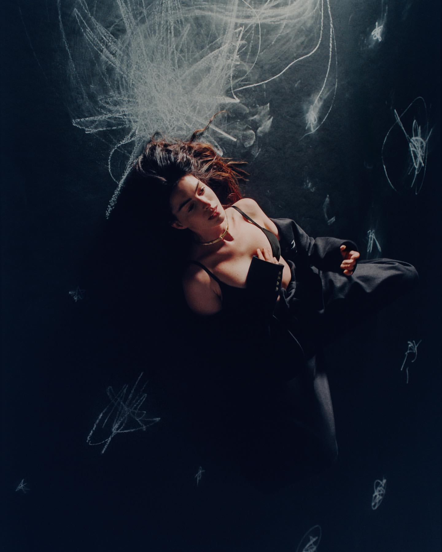 Fashionable brunette woman in black lingerie and jacket lying on her back surrounded by white chalk scribbles on a dark surface.
