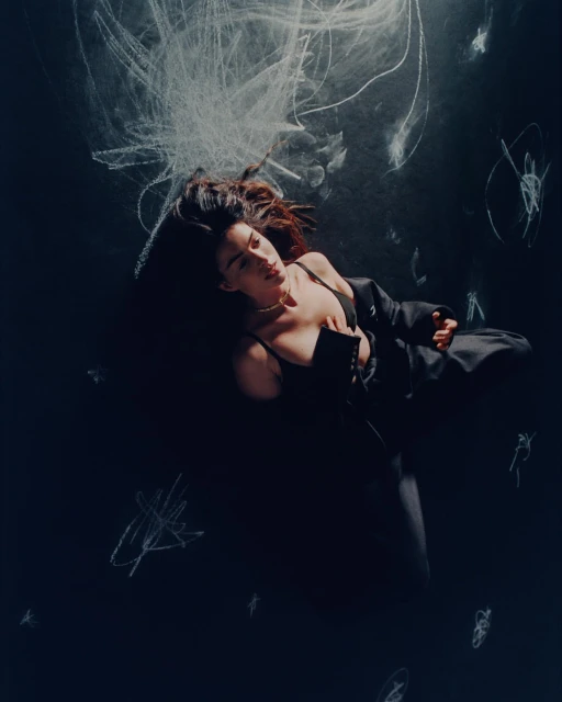 Fashionable brunette woman in black lingerie and jacket lying on her back surrounded by white chalk scribbles on a dark surface.