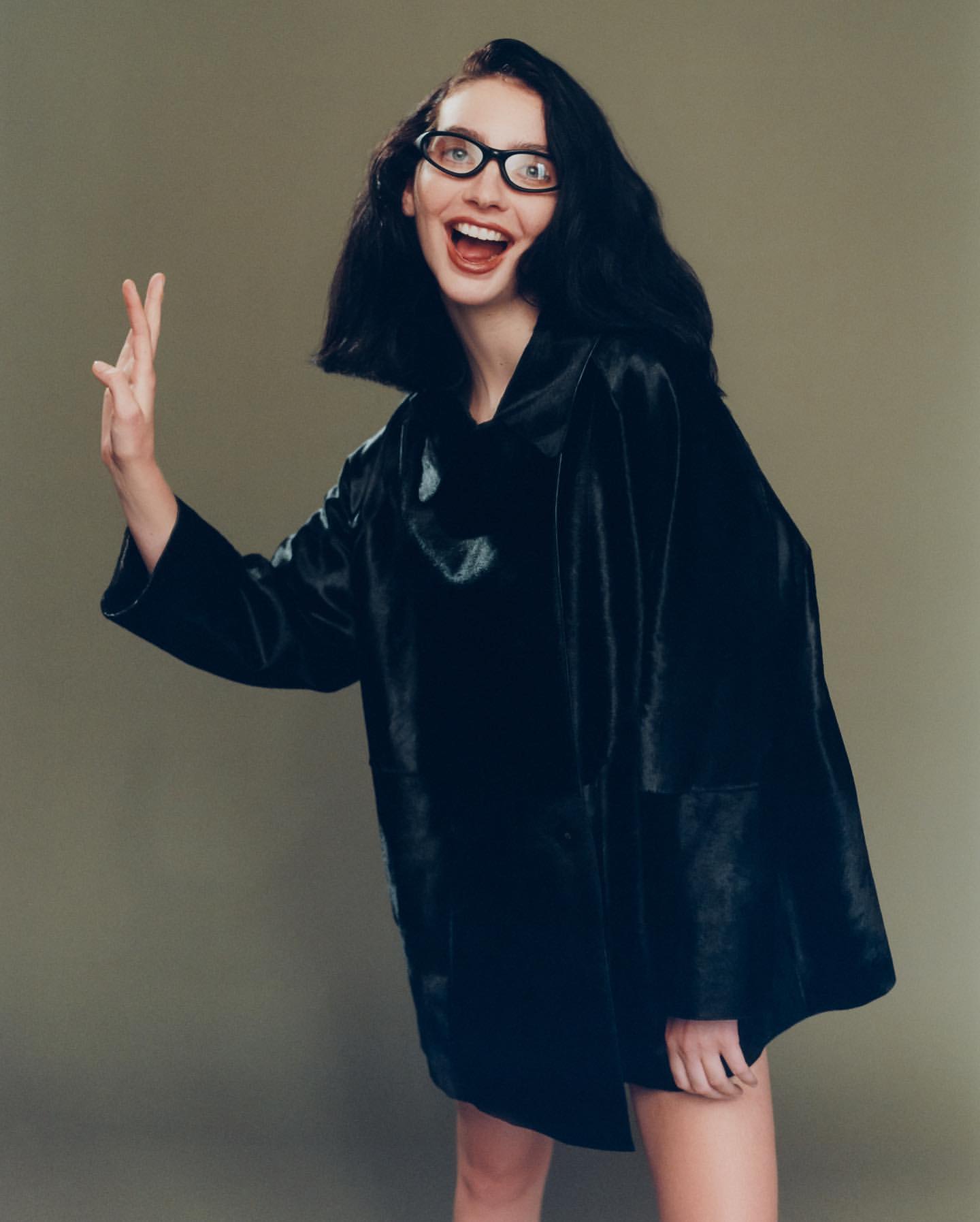 A young adult woman with dark hair wears black frame glasses and a dark, textured coat with a cheerful expression and an open mouth.