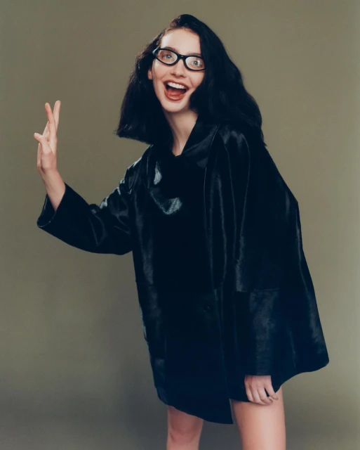 A young adult woman with dark hair wears black frame glasses and a dark, textured coat with a cheerful expression and an open mouth.