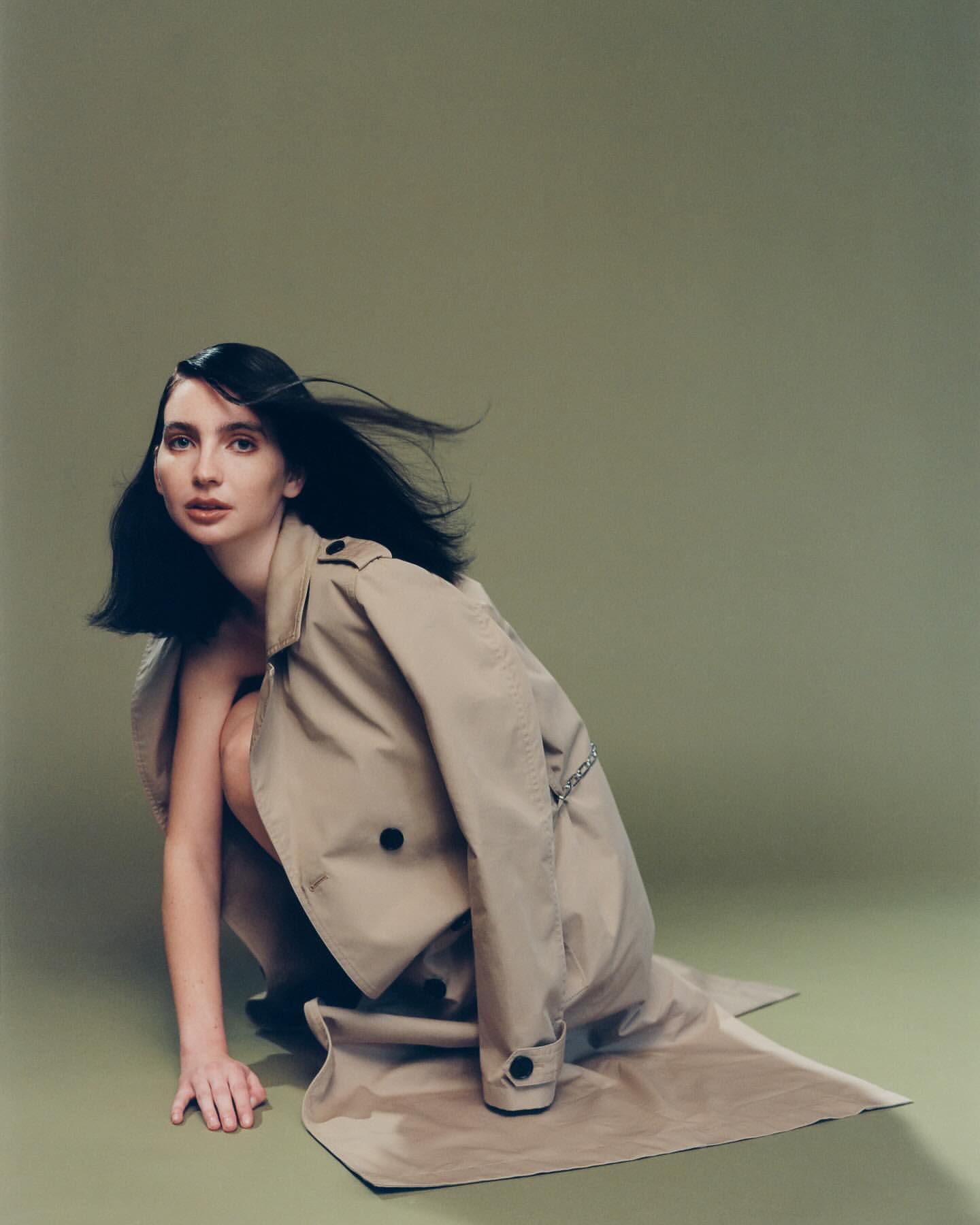 Portrait of a woman in a trench coat crouching against a sage green background in a fashion editorial style.
