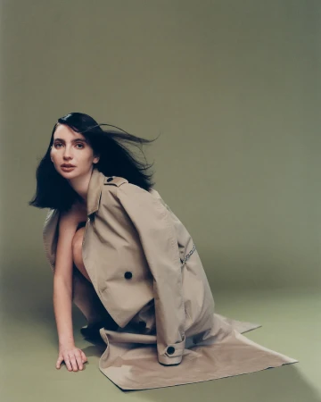 Portrait of a woman in a trench coat crouching against a sage green background in a fashion editorial style.