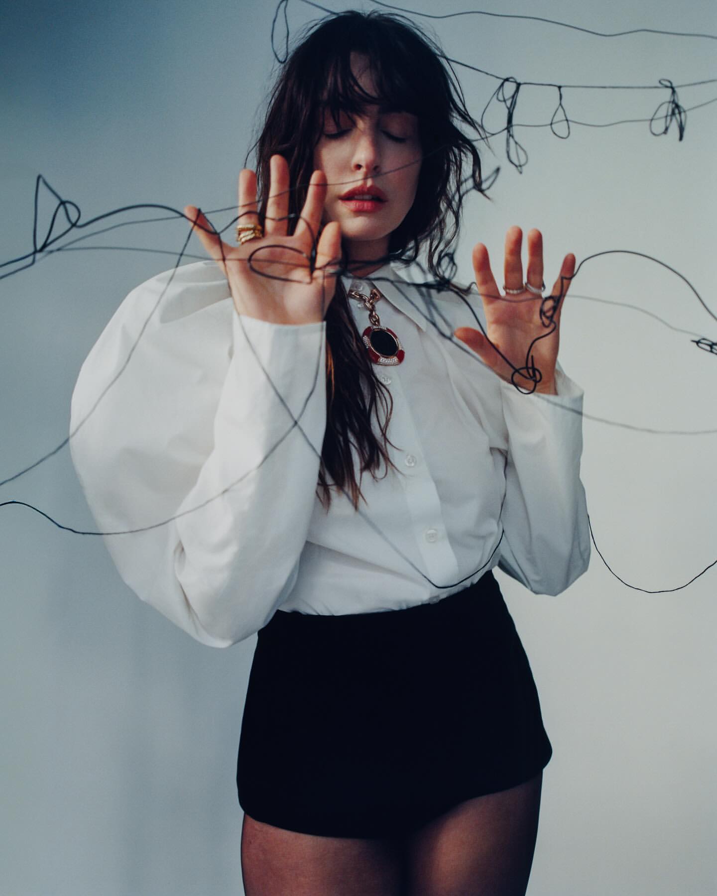 Woman in white puff-sleeve blouse and black shorts posing behind tangled black wire with eyes closed, photographed by Heather Hazzan.