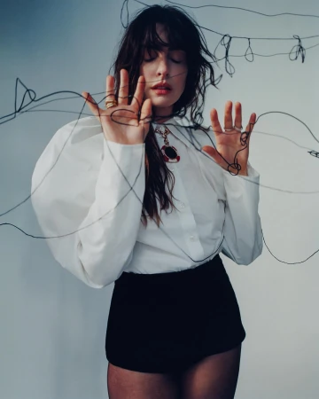 Woman in white puff-sleeve blouse and black shorts posing behind tangled black wire with eyes closed, photographed by Heather Hazzan.