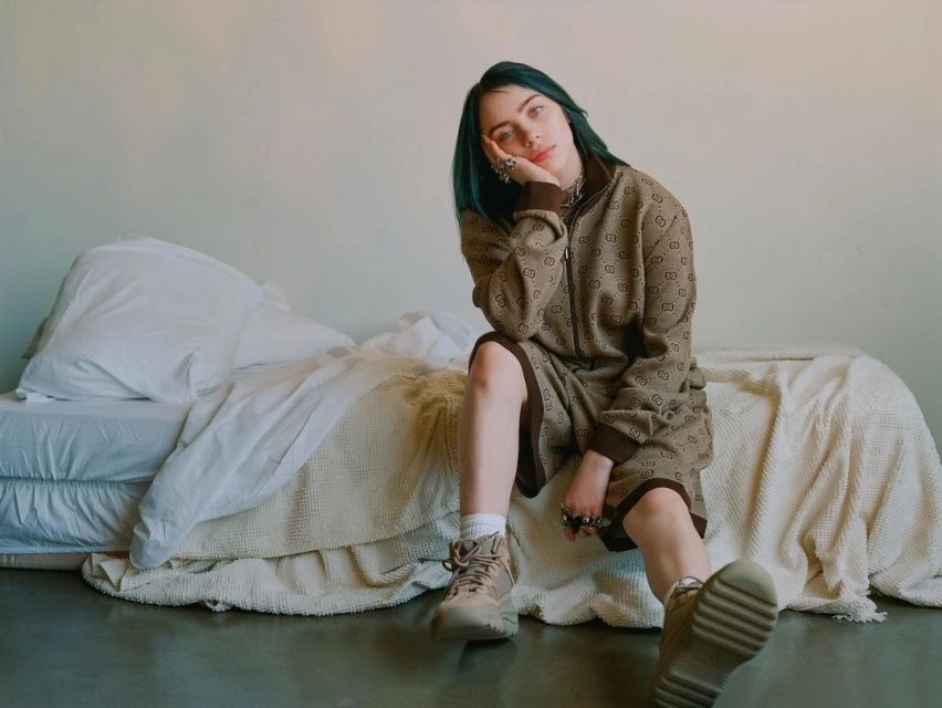 A young adult female with teal hair, wearing a brown patterned jacket and shorts, is sitting on a bed in an indoor setting.