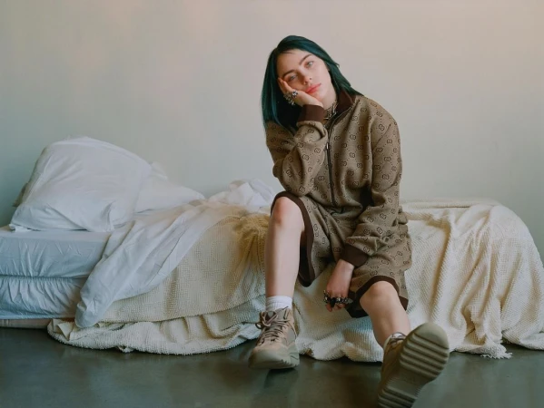 A young adult female with teal hair, wearing a brown patterned jacket and shorts, is sitting on a bed in an indoor setting.