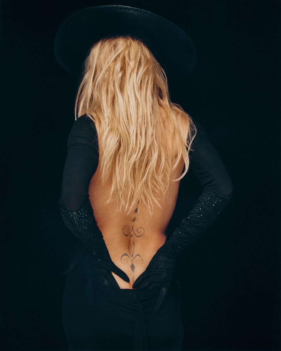 A woman with long blonde hair wears a black dress and hat, showing the tattoo on her back against a dark background in a studio shot.