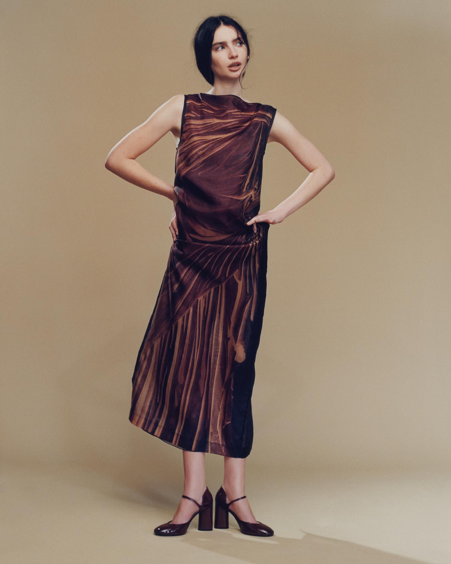 A slender woman poses in a sleeveless dress with brown patterns and chunky heeled shoes against a tan backdrop, fashion portrait.