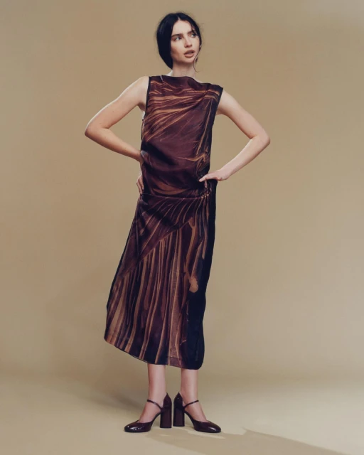 A slender woman poses in a sleeveless dress with brown patterns and chunky heeled shoes against a tan backdrop, fashion portrait.