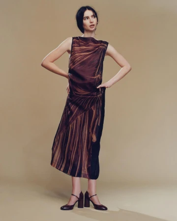 A slender woman poses in a sleeveless dress with brown patterns and chunky heeled shoes against a tan backdrop, fashion portrait.