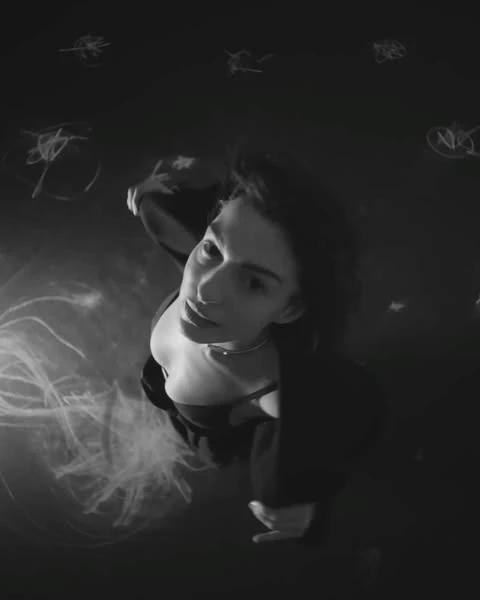 Portrait of an adult woman with dark hair looking up in a black and white image with dark clothing and abstract lighting effects.