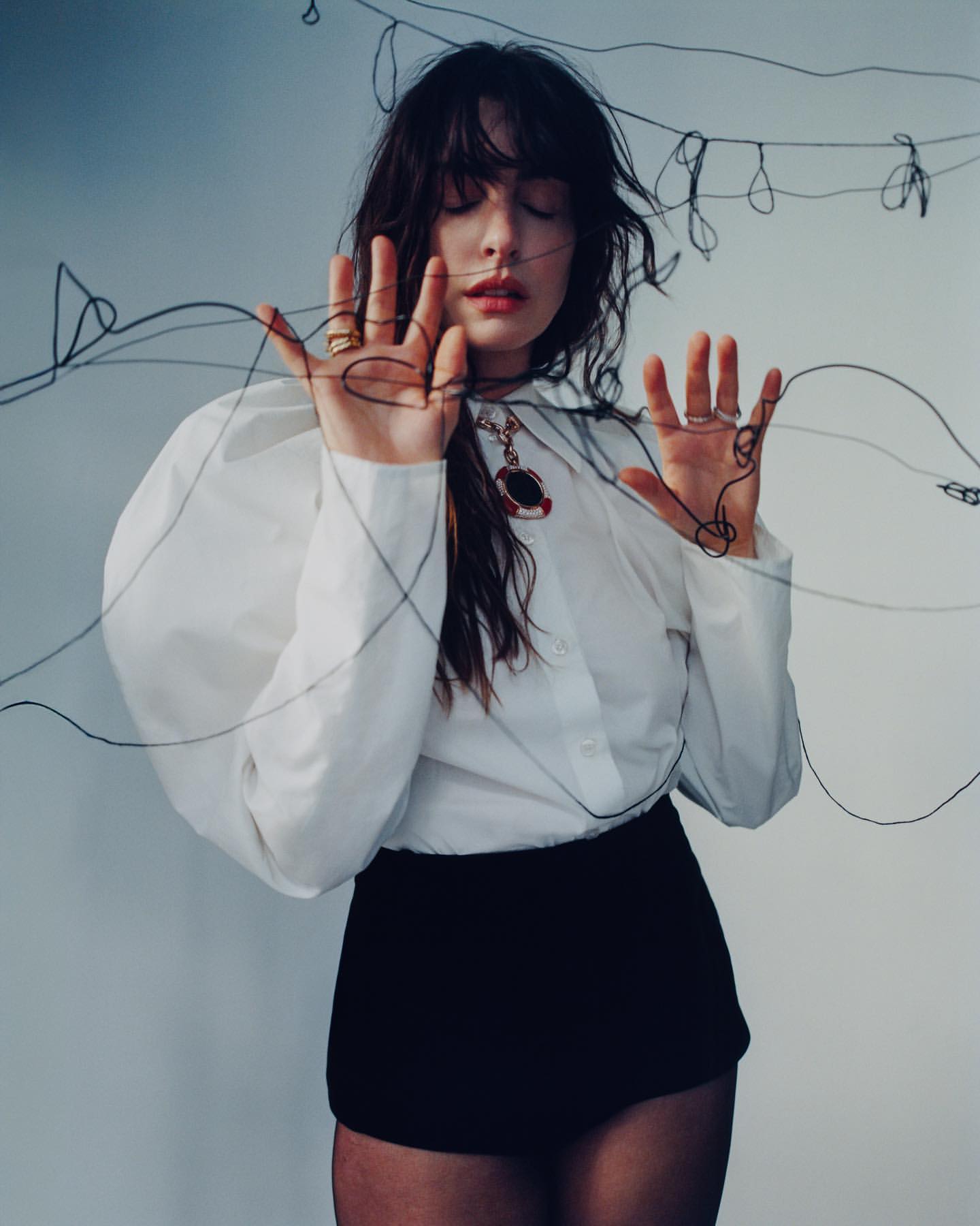 Portrait of a young woman with dark hair wearing a white blouse and black shorts, posing with black string against a soft white backdrop.