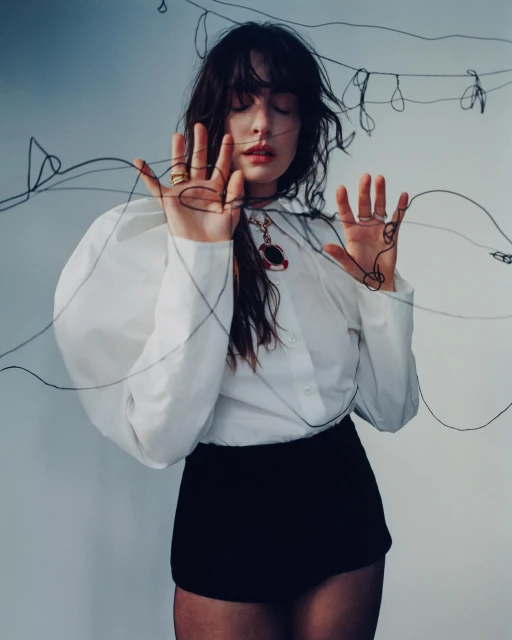 Portrait of a young woman with dark hair wearing a white blouse and black shorts, posing with black string against a soft white backdrop.