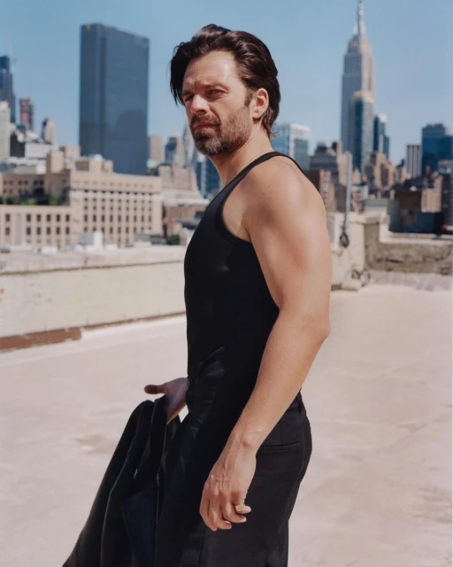 Portrait of actor Sebastian Stan in a black tank top and trousers on a rooftop in front of a city skyline with skyscrapers.
