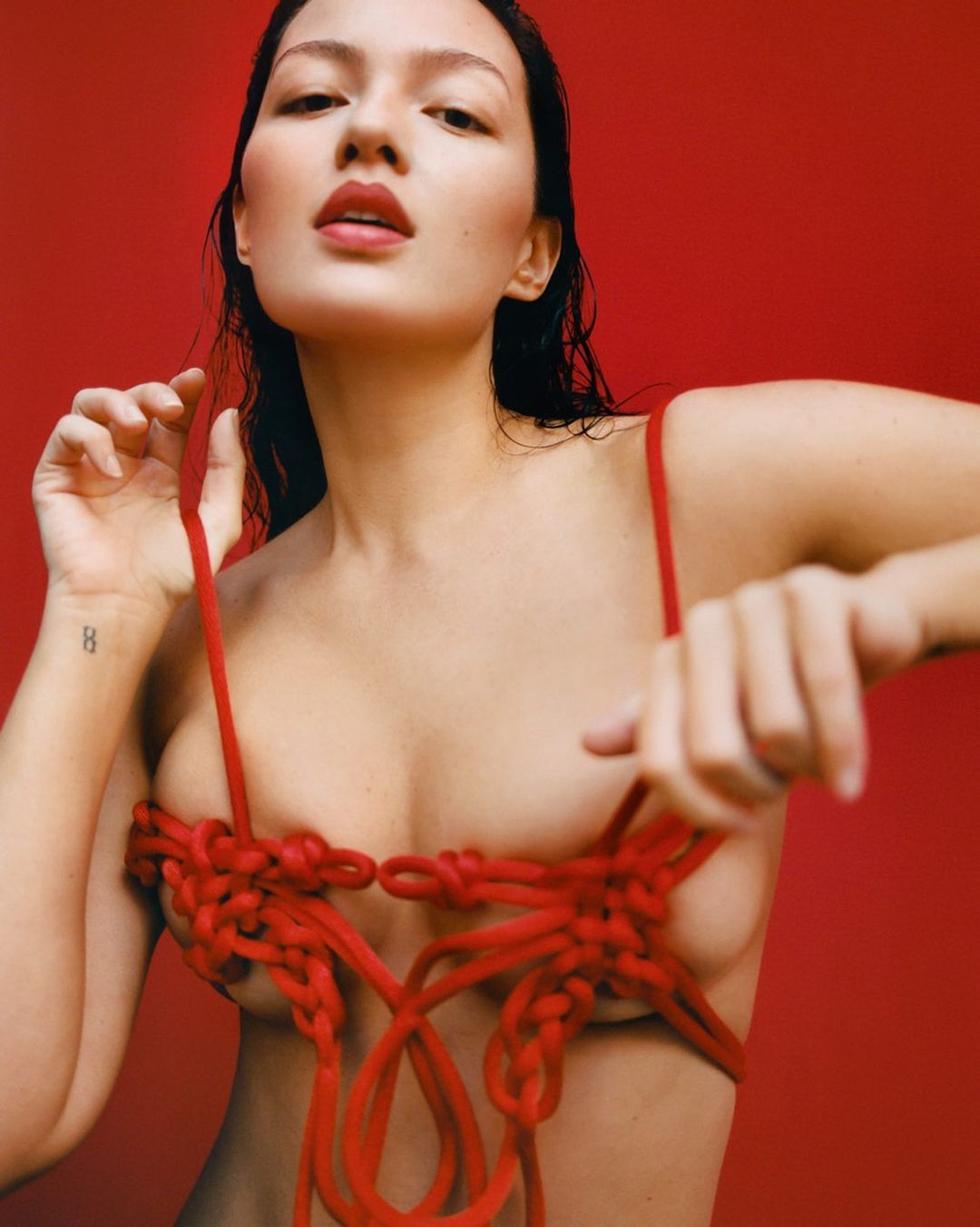 Portrait of a topless female model with red lipstick posing against a red background with rope art draped around her body.