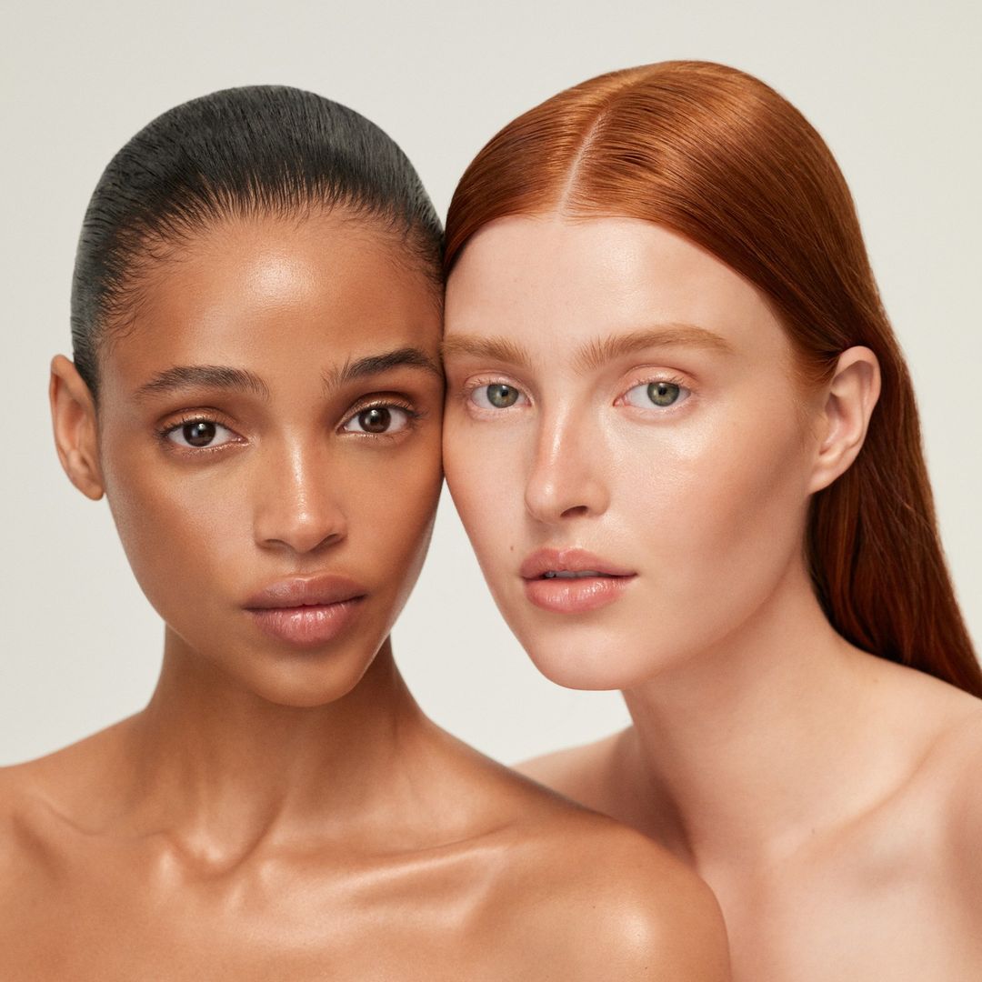 Close-up portrait of two young women with different skin tones and red and black hair, posing indoors against a neutral studio backdrop.