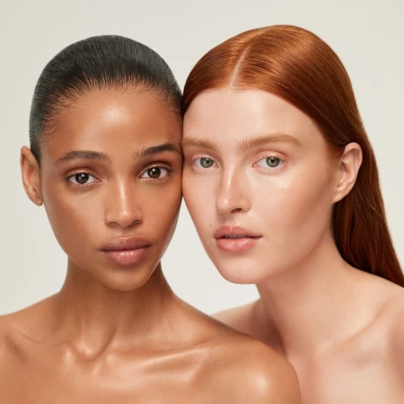 Close-up portrait of two women with glowing skin pressed cheek to cheek against a neutral light background