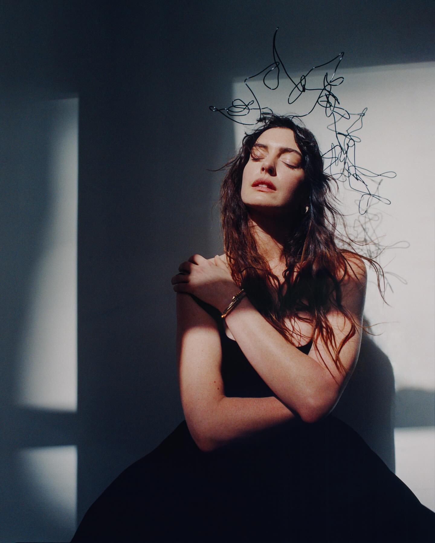 Portrait of a woman with her eyes closed, wearing a black dress and an elaborate wire headpiece, against a light gray wall with window shadows.