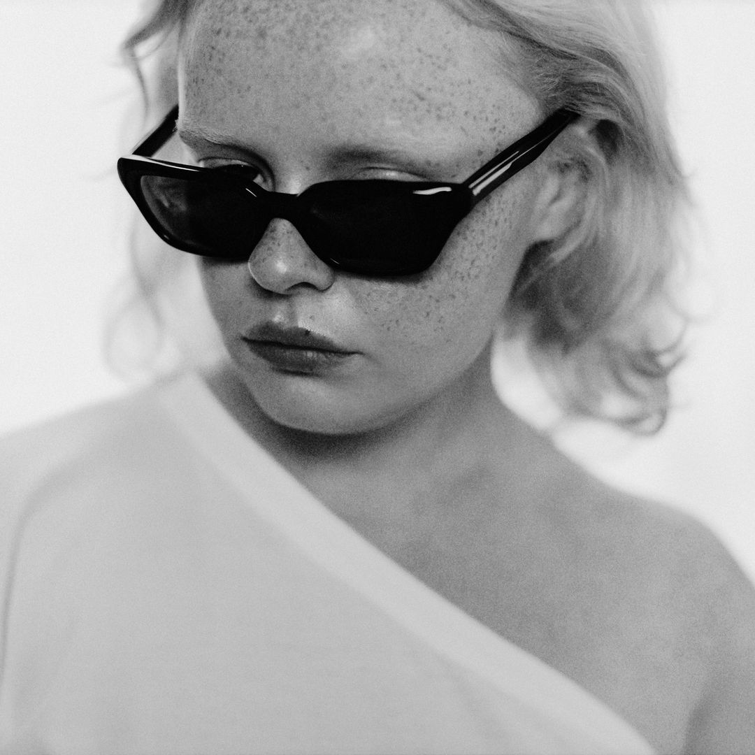 Close-up black and white portrait of a fair-skinned young woman wearing black angular sunglasses and a white top against a neutral backdrop.