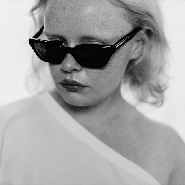 Close-up black and white portrait of a fair-skinned young woman wearing black angular sunglasses and a white top against a neutral backdrop.