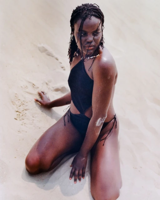Woman in a black swimsuit kneels in sand. She has dark hair and is wearing a beaded necklace. The background is light sand.