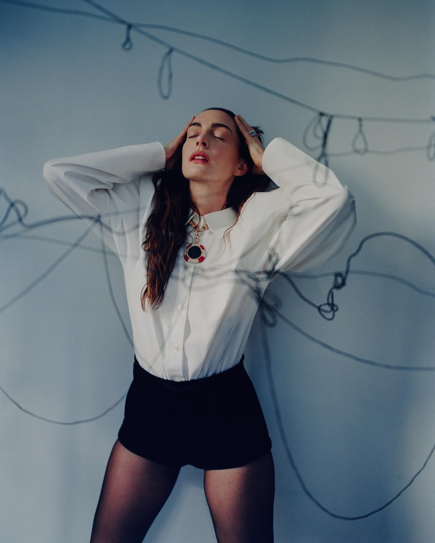 A woman with her eyes closed stands with her hands on her head. She is wearing a white shirt and dark shorts in a studio setting.