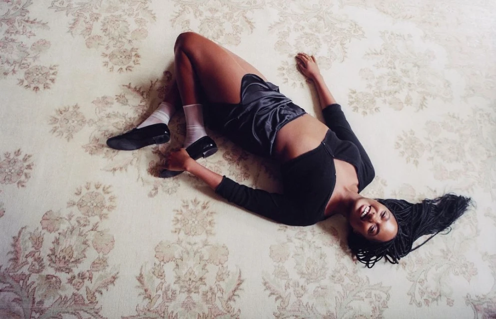 Smiling young woman lies on a floral rug wearing a black top, satin shorts, socks, and black tap shoes with braided hair.