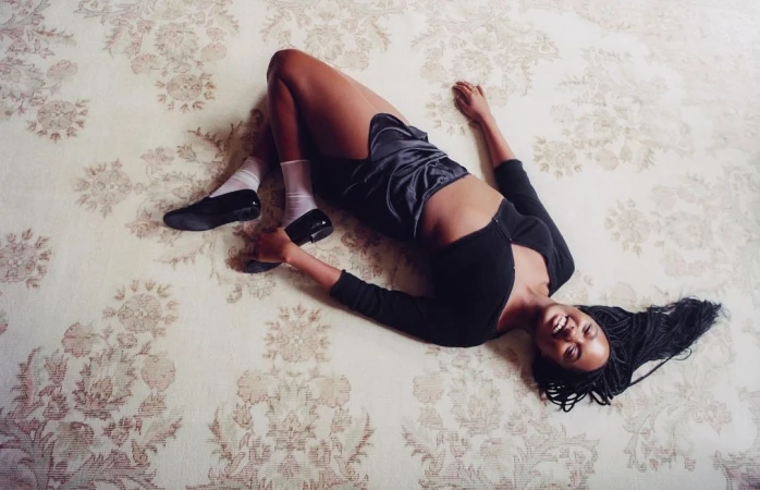 Smiling young woman lies on a floral rug wearing a black top, satin shorts, socks, and black tap shoes with braided hair.
